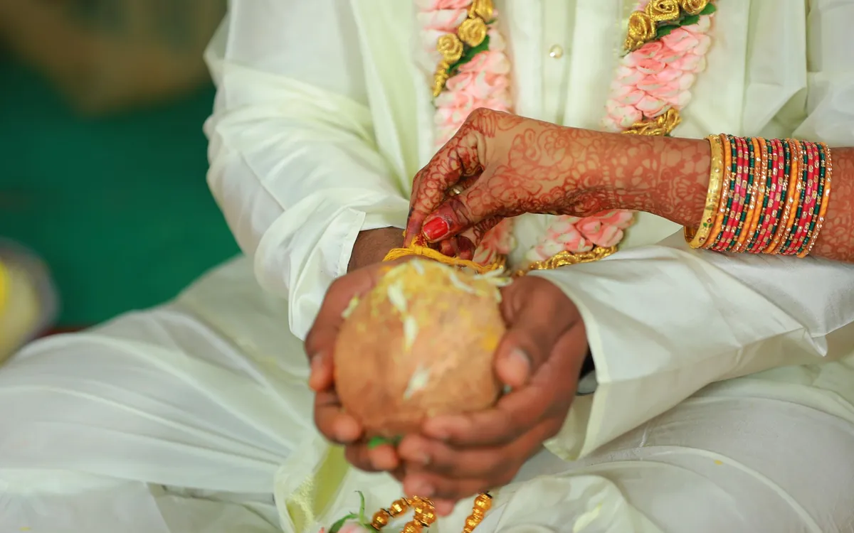 A groom's hands holding a coconut while the bride touches it